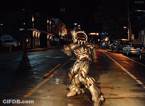 A Man In A Robot Costume Is Walking Down A Street At Night In Front Of A Cafe Sign GIF