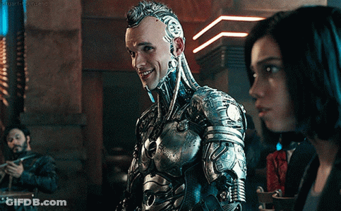 A Man In A Robotic Suit Stands Next To A Woman In A Dark Room GIF