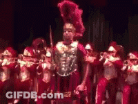 A Man In A Roman Costume Is Standing In Front Of A Marching Band Holding A Sword . GIF