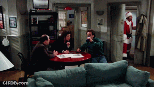 A Man In A Santa Suit Stands In A Living Room With A Group Of People Sitting Around A Table GIF