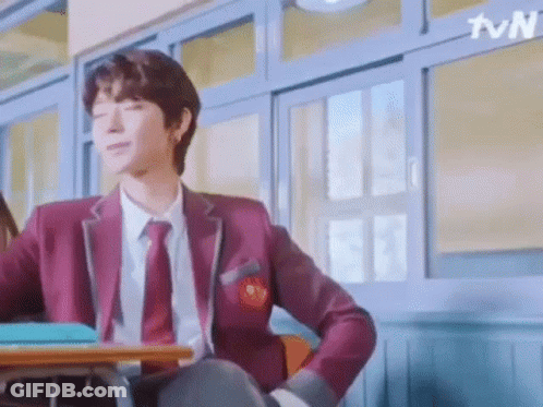 A Man In A School Uniform Is Sitting At A Desk In A Classroom . GIF