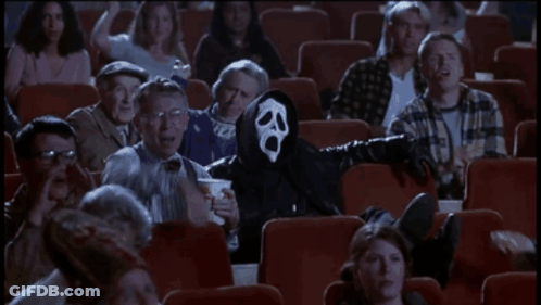 A Man In A Scream Mask Is Sitting In A Theater With Other People GIF