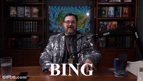A Man In A Sequined Jacket Is Sitting At A Desk With The Word Bing Written On It GIF