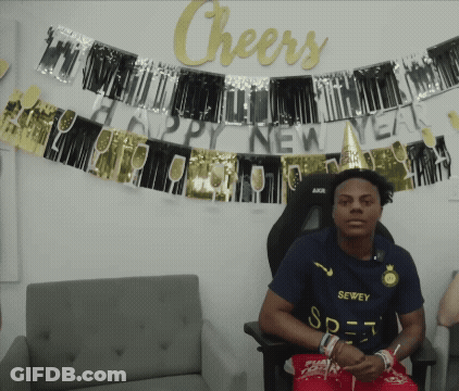 A Man In A Sewee Shirt Sits In Front Of A Sign That Says Cheers GIF