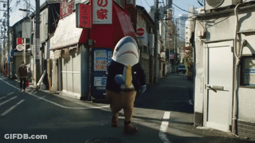 A Man In A Shark Costume Walks Down A Street In Front Of A Store With Chinese Writing On It GIF