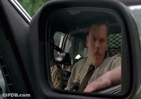 A Man In A Sheriff 'S Uniform Is Driving A Car And Looking In The Rear View Mirror . GIF