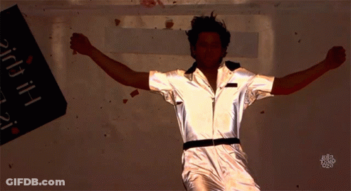 A Man In A Silver Jumpsuit Is Dancing In Front Of A Sign That Says ' Bitch ' GIF