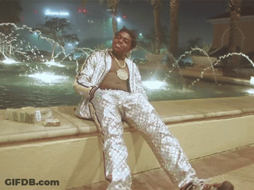 A Man In A Silver Suit Sits In Front Of A Fountain With A Sign That Says Green Juice GIF