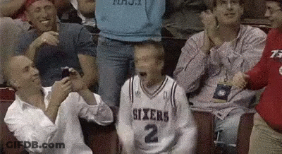 A Man In A Sixers Jersey Is Sitting In A Crowd Of People Watching A Basketball Game . GIF