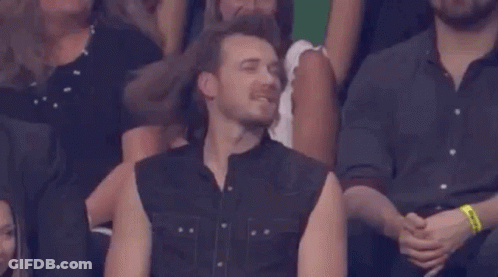 A Man In A Sleeveless Shirt Is Sitting In A Crowd GIF