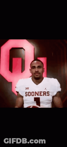 A Man In A Sooners Jersey Is Holding A Football In Front Of A Large Red Q . GIF