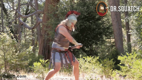 A Man In A Spartan Costume Is Holding A Sword In The Woods With A Dr.squatch Logo Behind Him GIF