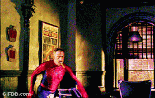 A Man In A Spiderman Costume Is Standing In A Room Next To A Sign That Says Wanted . GIF