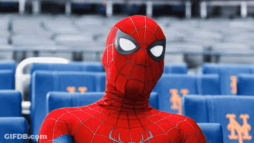 A Man In A Spiderman Costume Sits In A Stadium With Blue Seats With Ny On Them GIF