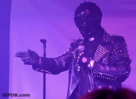 A Man In A Spiked Leather Jacket Is Singing Into A Microphone In A Dark Room GIF
