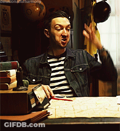 A Man In A Striped Shirt And Denim Jacket Is Sitting At A Desk GIF