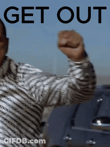 A Man In A Striped Shirt Holds Up His Fist In Front Of A Sign That Says Get Out GIF