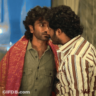 A Man In A Striped Shirt Is Kissing Another Man In A Green Shirt GIF