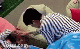 A Man In A Striped Shirt Is Putting His Hand On Another Man 'S Forehead While Laying On A Bed . GIF
