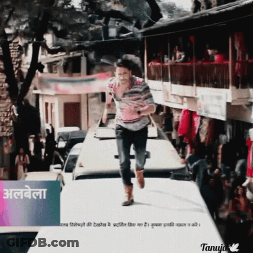 A Man In A Striped Shirt Is Running Down A Street In Front Of A Sign That Says Tanuja GIF