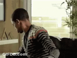 A Man In A Striped Shirt Is Sitting On A Couch Looking Out A Window . GIF