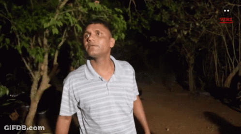 A Man In A Striped Shirt Is Standing In Front Of Trees At Night . GIF