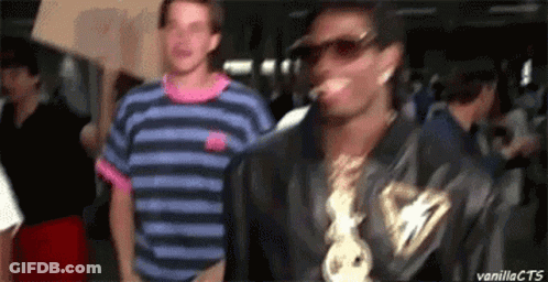 A Man In A Striped Shirt Is Standing Next To A Man Wearing Sunglasses And A Chain Around His Neck . GIF