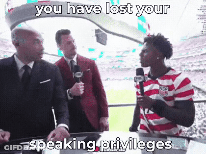 A Man In A Striped Shirt Is Talking Into A Microphone While Two Men In Suits Look On . GIF