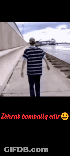 A Man In A Striped Shirt Is Walking Down A Sidewalk Next To The Water . GIF