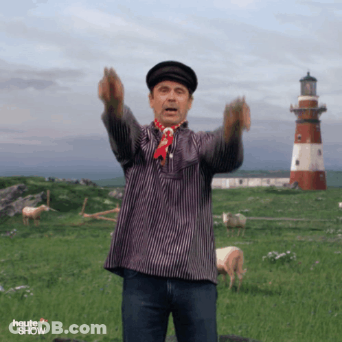 A Man In A Striped Shirt Stands In A Field With A Lighthouse In The Background And The Word Heute Show On The Bottom GIF