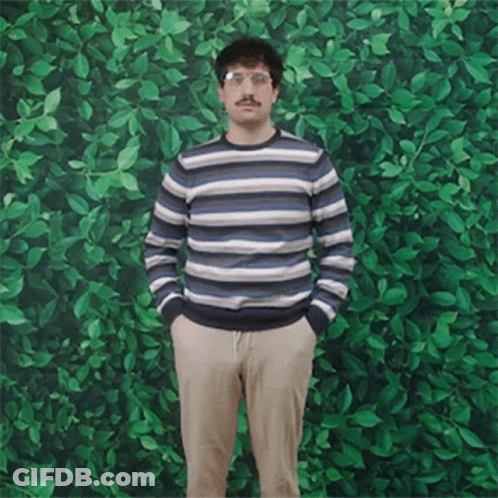 A Man In A Striped Sweater Stands With His Hands In His Pockets In Front Of A Green Wall GIF