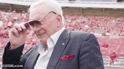 A Man In A Suit Adjusts His Sunglasses In Front Of A Crowd In A Stadium GIF
