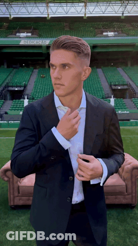 A Man In A Suit Adjusts His Tie In Front Of A Stadium That Says Dedicacao On It GIF