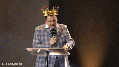 A Man In A Suit And A Crown Is Standing At A Podium With A Microphone . GIF