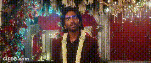 A Man In A Suit And A Garland Around His Neck Is Standing In Front Of A Red Curtain . GIF