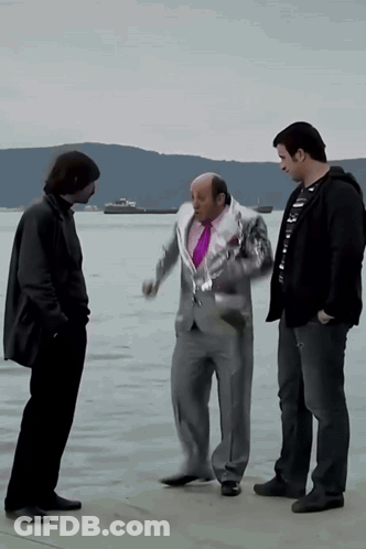 A Man In A Suit And A Purple Tie Talks To Two Other Men In Front Of A Body Of Water GIF