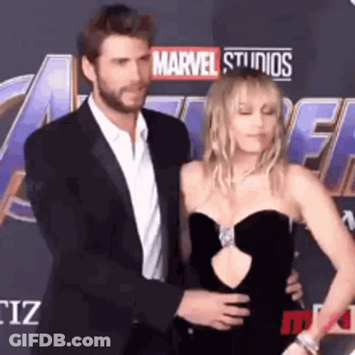 A Man In A Suit And A Woman In A Black Dress Are Standing On A Red Carpet . GIF