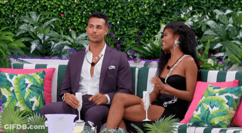 A Man In A Suit And A Woman In A Black Dress Sit On A Couch GIF