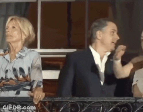 A Man In A Suit And A Woman In A Floral Shirt Are Standing On A Balcony GIF