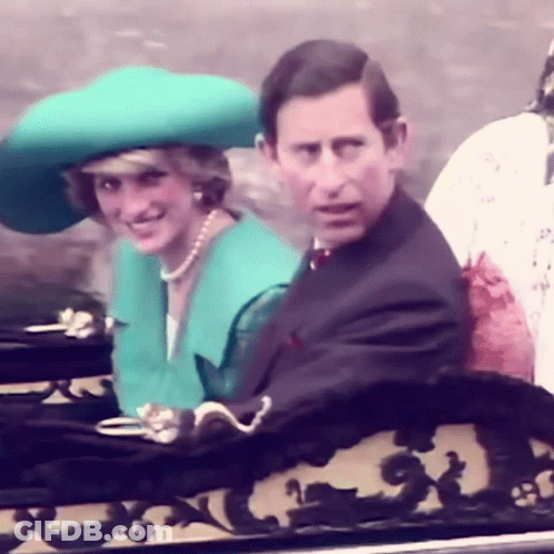 A Man In A Suit And A Woman In A Green Hat Are Sitting Next To Each Other On A Balcony . GIF