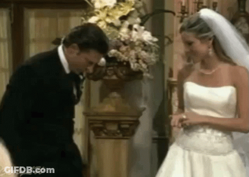 A Man In A Suit And A Woman In A Wedding Dress Are Standing Next To Each Other In Front Of A Vase Of Flowers GIF