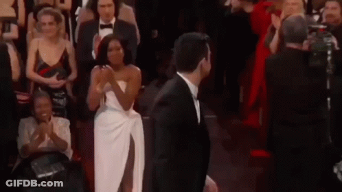 A Man In A Suit And A Woman In A White Dress Are Standing In Front Of A Crowd Of People . GIF