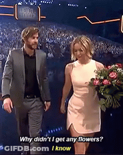 A Man In A Suit And A Woman In A White Dress Holding A Bouquet Of Flowers Are Walking On A Stage . GIF