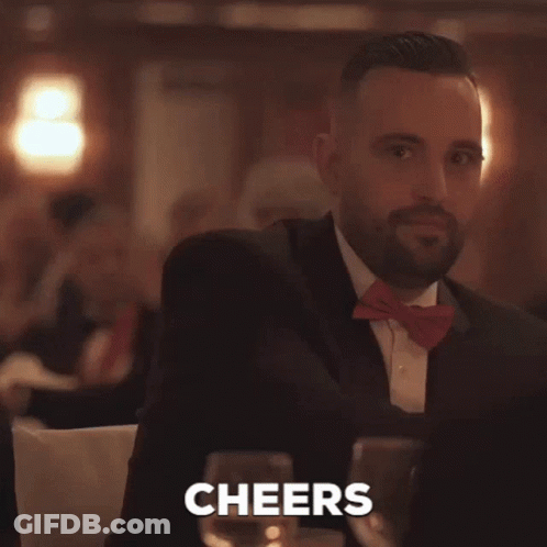 A Man In A Suit And Bow Tie Is Holding A Glass Of Wine And The Word Cheers Is Behind Him GIF