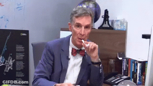 A Man In A Suit And Bow Tie Is Sitting At A Desk In Front Of A National Geographic Sign GIF