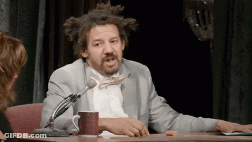 A Man In A Suit And Bow Tie Is Sitting At A Table With A Microphone And A Cup Of Coffee . GIF
