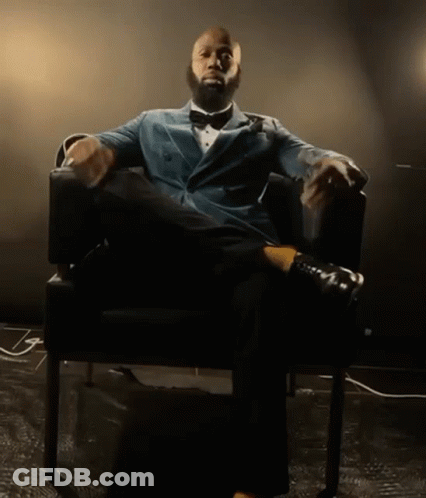 A Man In A Suit And Bow Tie Is Sitting In A Chair With His Legs Crossed GIF