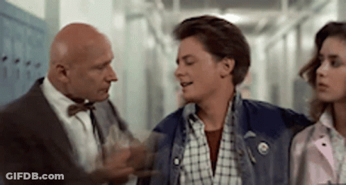 A Man In A Suit And Bow Tie Is Talking To A Young Man In A Plaid Shirt In A Hallway . GIF