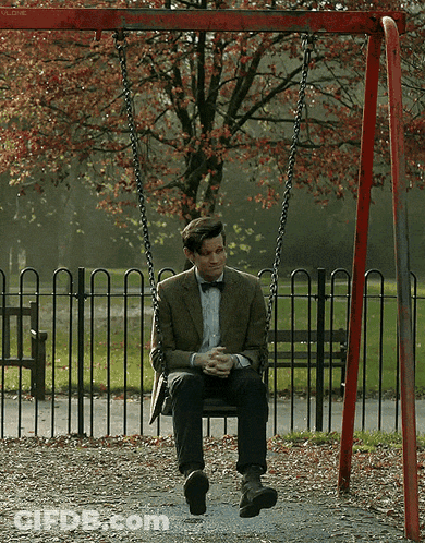 A Man In A Suit And Bow Tie Sits On A Swing In A Park GIF
