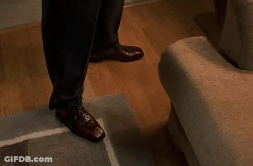 A Man In A Suit And Brown Shoes Is Walking Down Stairs In A Living Room . GIF